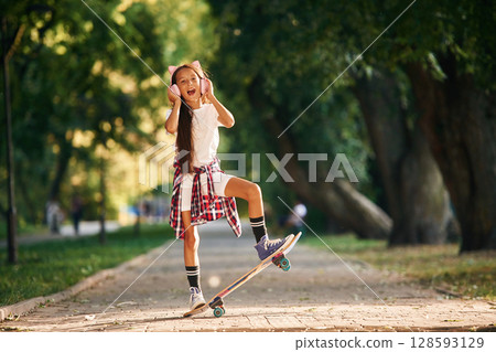 Listening to the music in cute headphones. Happy little girl with skateboard outdoors Listening to the music in cute headphones. Happy little girl with skateboard outdoors 128593129