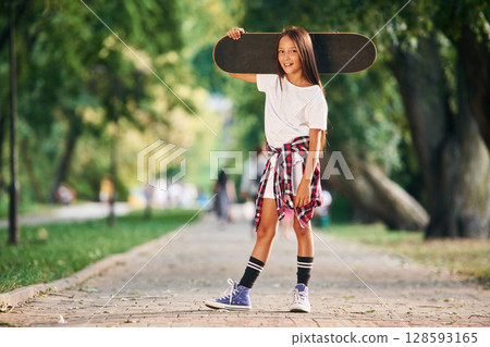 Holding the skateboard in the hand, on the shoulder. Happy little girl is outdoors Holding the skateboard in the hand, on the shoulder. Happy little girl is outdoors 128593165