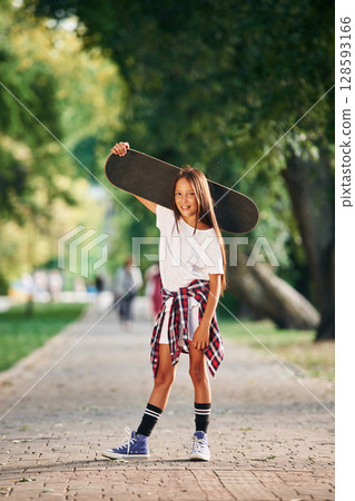 Holding the skateboard in the hand, on the shoulder. Happy little girl is outdoors Holding the skateboard in the hand, on the shoulder. Happy little girl is outdoors 128593166