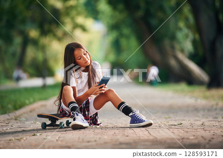 Blue smartphone is in hands. Happy little girl with skateboard outdoors 128593171