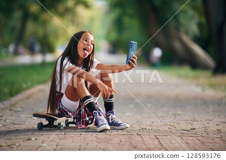 Showing tongue, making selfie. Happy little girl with skateboard outdoors Showing tongue, making selfie. Happy little girl with skateboard outdoors 128593176