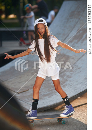 Riding skateboard near the ramp. Happy little girl with is outdoors Riding skateboard near the ramp. Happy little girl with is outdoors 128593215
