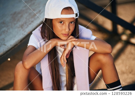 Sitting and looking at the camera. Young girl in casual clothes is in the public park outdoors 128593226