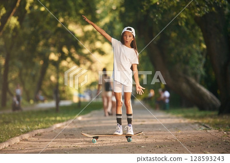 Standing in the public park. Happy little girl with skateboard outdoors Standing in the public park. Happy little girl with skateboard outdoors 128593243