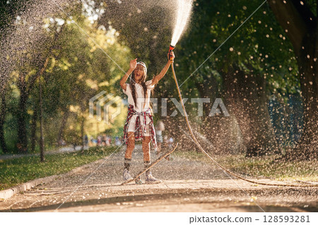 Pouring water from a hose. Happy little girl with skateboard outdoors Pouring water from a hose. Happy little girl with skateboard outdoors 128593281