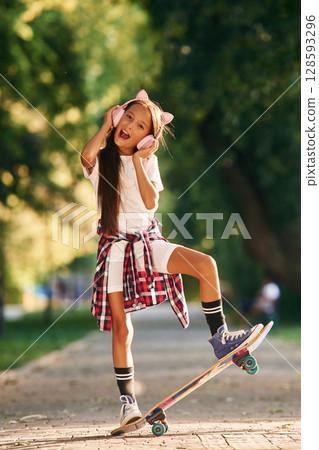 Listening to the music in cute headphones. Happy little girl with skateboard outdoors 128593296
