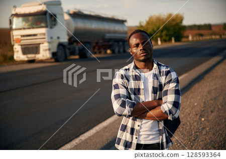 Serious look. With arms crossed. Black man is standing on the road with truck on it Serious look. With arms crossed. Black man is standing on the road with truck on it 128593364