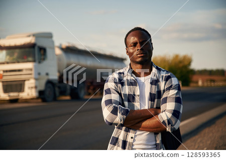 Serious look. With arms crossed. Black man is standing on the road with truck on it Serious look. With arms crossed. Black man is standing on the road with truck on it 128593365