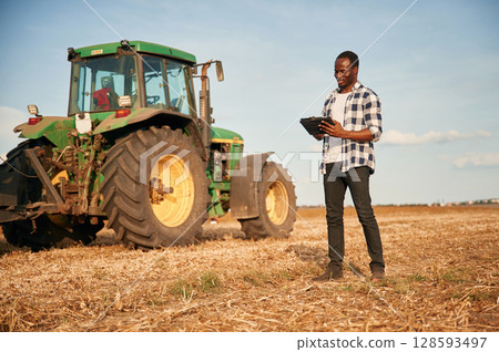 The tractor is parked behind. Beautiful African American man is in the agricultural field The tractor is parked behind. Beautiful African American man is in the agricultural field 128593497