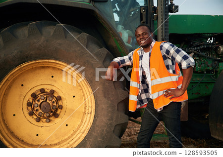 Leaning on the big wheel of tractor. Beautiful African American man is in the agricultural field Leaning on the big wheel of tractor. Beautiful African American man is in the agricultural field 128593505