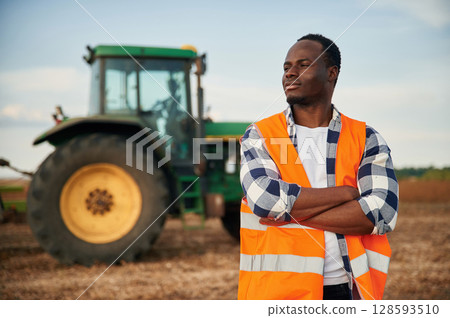 Near the tractor in orange uniform. Beautiful African American man is in the agricultural field Near the tractor in orange uniform. Beautiful African American man is in the agricultural field 128593510