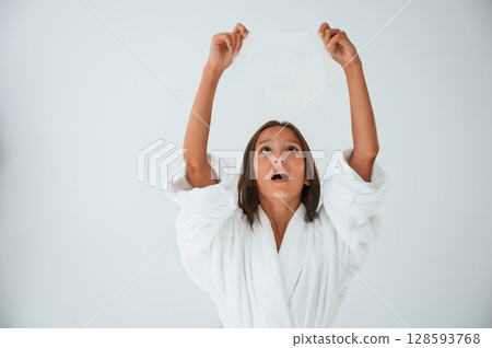 Standing, having fun, in white robe, holding mask. Conception of beauty and self care. Young girl is in the studio Standing, having fun, in white robe, holding mask. Conception of beauty and self care. Young girl is in the studio 128593768