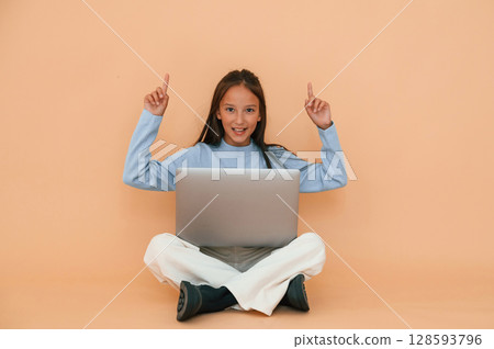 Pointing to the top. Sitting with laptop. Cute young girl is in the studio against background Pointing to the top. Sitting with laptop. Cute young girl is in the studio against background 128593796