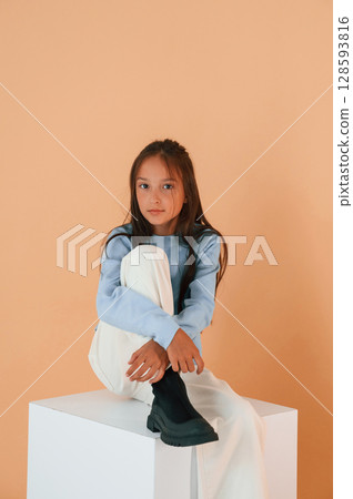Sitting on white cube. Cute young girl is in the studio against background 128593816
