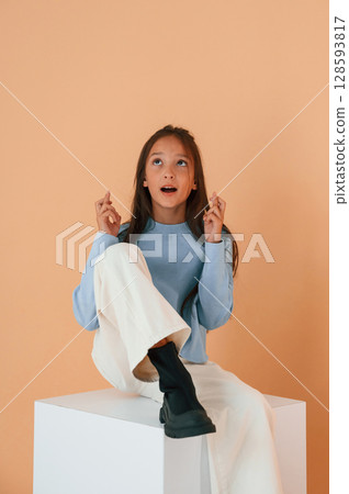 Sitting on cube with fingers crossed. Cute young girl is in the studio against background 128593817