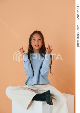 Sitting on cube with fingers crossed. Cute young girl is in the studio against background 128593818