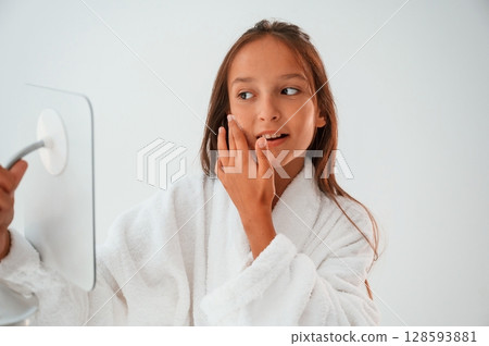 Using cream on face, holding mirror. Conception of beauty and self care. Young girl is in the studio 128593881