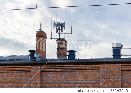 An old air raid warning siren stands atop a city building rooftop with classic brick chimneys, symbolizing crucial public safety and emergency readiness systems An old air raid warning siren stands atop a city building rooftop with classic brick chimneys, symbolizing crucial public safety and emergency readiness systems 128593943