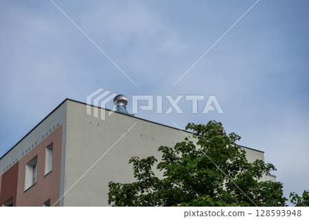 Modern building corner showcasing a functional rooftop ventilation system and lush green tree against a clear sky, highlighting sustainable urban infrastructure development Modern building corner showcasing a functional rooftop ventilation system and lush green tree against a clear sky, highlighting sustainable urban infrastructure development 128593948