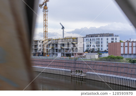 Urban construction scene featuring a modern residential building under development with cranes, alongside a river and its brick embankment Urban construction scene featuring a modern residential building under development with cranes, alongside a river and its brick embankment 128593970