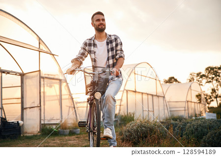 Front view. Handsome man in casual clothes is with bicycle on the agricultural field near greenhouse 128594189