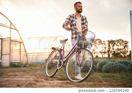 Front view. Handsome man in casual clothes is with bicycle on the agricultural field near greenhouse 128594190