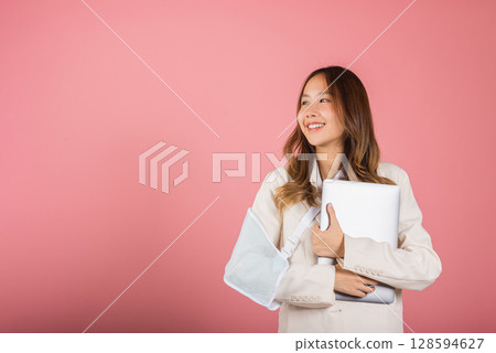 Asian business woman confident smiling broken arm after accident and wear arm splint for treatment but still have to work she hold laptop computer studio isolated on pink background,copy space 128594627
