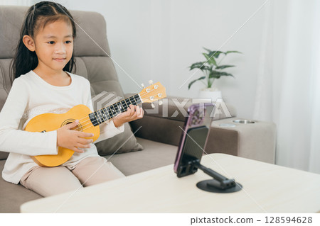 A young girl learns music on a ukulele, sitting on a couch while a smartphone streams her session live. The cozy home atmosphere enhances the focus on creative online education. 128594628