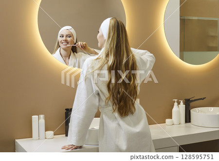 Young woman with long hair in bathrobe brushing teeth with toothbrush Young woman with long hair in bathrobe brushing teeth with toothbrush 128595751