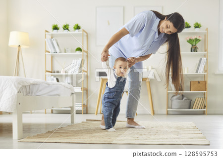 Childs first steps in learning to walk at home, mother holding hands of toddler with love and care Childs first steps in learning to walk at home, mother holding hands of toddler with love and care 128595753