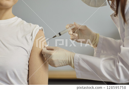 Injection into patient arm, medical professional wearing sterile gloves, white coat, holding syringe 128595758
