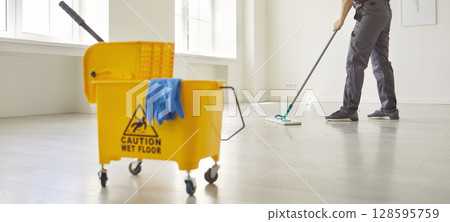 Janitor with mop cleaning empty office floor, worker mopping by cart with supplies and warning sign 128595759