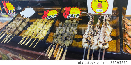 Grilled street food skewers in Thailand, including chicken satay, beef, octopus, and seafood, displayed on a market counter with price signs and vibrant labels. Grilled street food skewers in Thailand, including chicken satay, beef, octopus, and seafood, displayed on a market counter with price signs and vibrant labels. 128595881