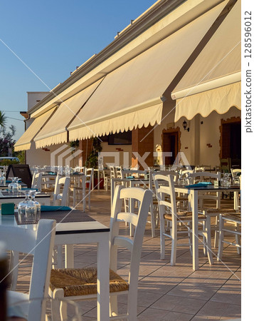 Empty outdoor restaurant terrace in Sardinia, Italy, bathed in golden evening sunlight with white chairs and covered awning. Empty outdoor restaurant terrace in Sardinia, Italy, bathed in golden evening sunlight with white chairs and covered awning. 128596012