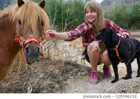 Woman and Black Dog Engaging with a Miniature Horse in a Scenic Outdoor Setting Woman and Black Dog Engaging with a Miniature Horse in a Scenic Outdoor Setting 128596158