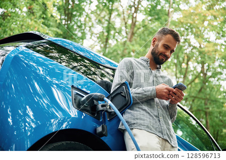 View from below, with smartphone. Handsome man is standing outdoors near electric automobile and waiting for charging View from below, with smartphone. Handsome man is standing outdoors near electric automobile and waiting for charging 128596713