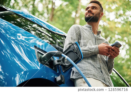 View from below, with smartphone. Handsome man is standing outdoors near electric automobile and waiting for charging View from below, with smartphone. Handsome man is standing outdoors near electric automobile and waiting for charging 128596716