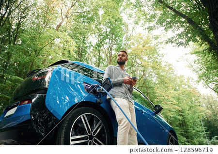 Trees behind. Handsome man is standing outdoors near electric automobile and waiting for charging Trees behind. Handsome man is standing outdoors near electric automobile and waiting for charging 128596719