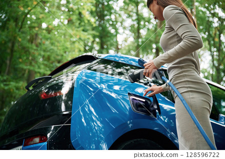 Young woman is putting charger into the socket of electric automobile Young woman is putting charger into the socket of electric automobile 128596722