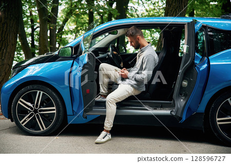With smartphone in hands. Beautiful young man is sitting in the blue car outdoors With smartphone in hands. Beautiful young man is sitting in the blue car outdoors 128596727