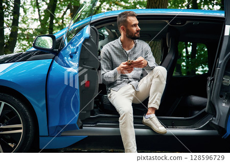 With smartphone in hands. Beautiful young man is sitting in the blue car outdoors 128596729