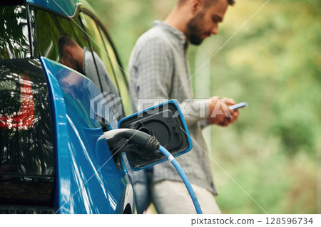 Trees behind. Handsome man is standing outdoors near electric automobile and waiting for charging 128596734