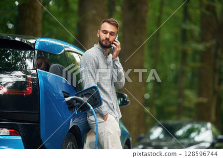 Wireless device, using smartphone. Handsome man is standing outdoors near electric automobile and waiting for charging 128596744