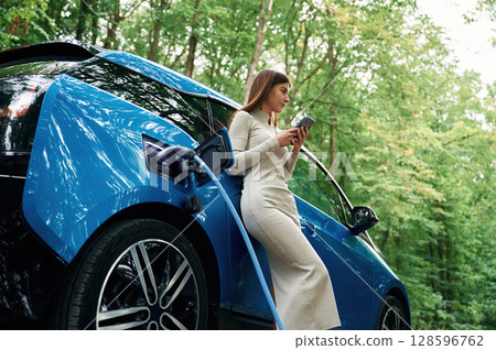 Leaning, using smartphone. Young woman is standing outdoors near electric automobile that is charging 128596762