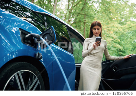 Leaning, using smartphone. Young woman is standing outdoors near electric automobile that is charging Leaning, using smartphone. Young woman is standing outdoors near electric automobile that is charging 128596766