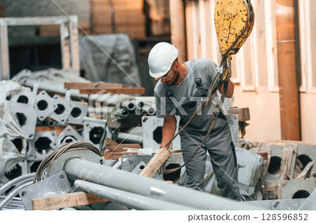 Using crane hook. Young factory worker in grey uniform Using crane hook. Young factory worker in grey uniform 128596852