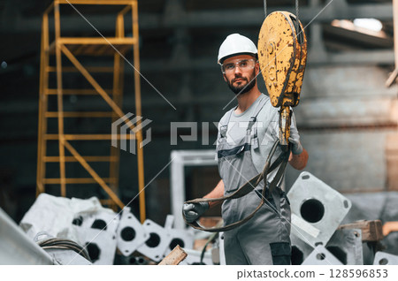 Using crane hook. Young factory worker in grey uniform Using crane hook. Young factory worker in grey uniform 128596853