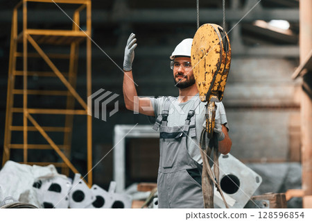 Using crane hook. Young factory worker in grey uniform Using crane hook. Young factory worker in grey uniform 128596854