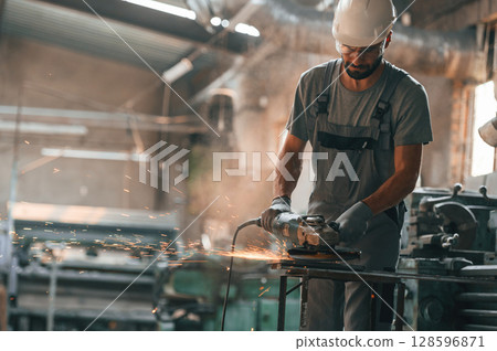 Angle grinder in hands. Young factory worker in grey uniform Angle grinder in hands. Young factory worker in grey uniform 128596871