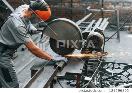 Focused at work, cutting the metal. Young man at factory in grey uniform 128596895